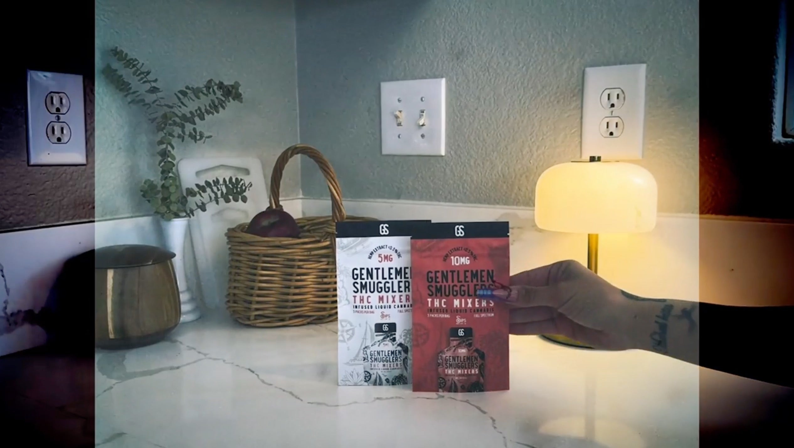 Hand holding Gentlemen Smugglers THC Mixer pouches on a marble kitchen counter with a basket, plant, and lamp nearby.